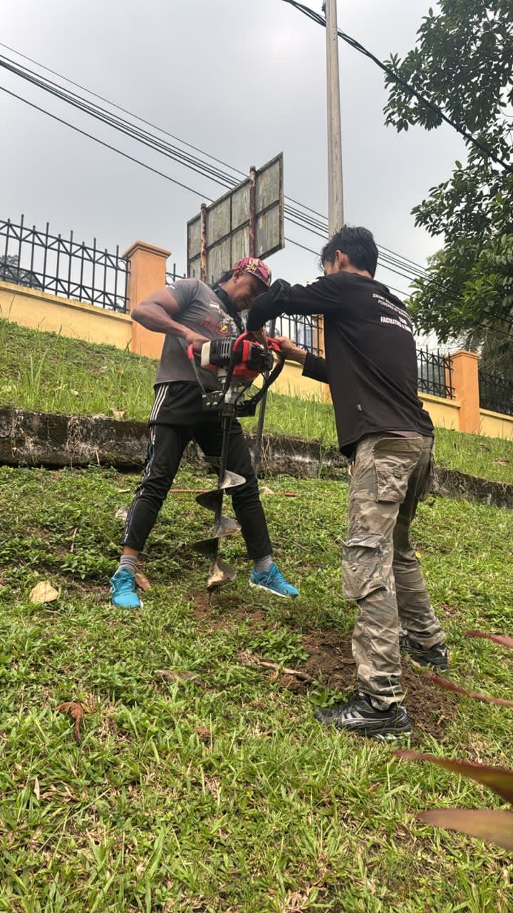 Two workers drilling a foundation hole with a petrol auger on the grassy slope