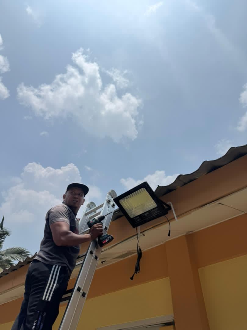 A worker on a ladder mounting a solar floodlight under the roof eaves